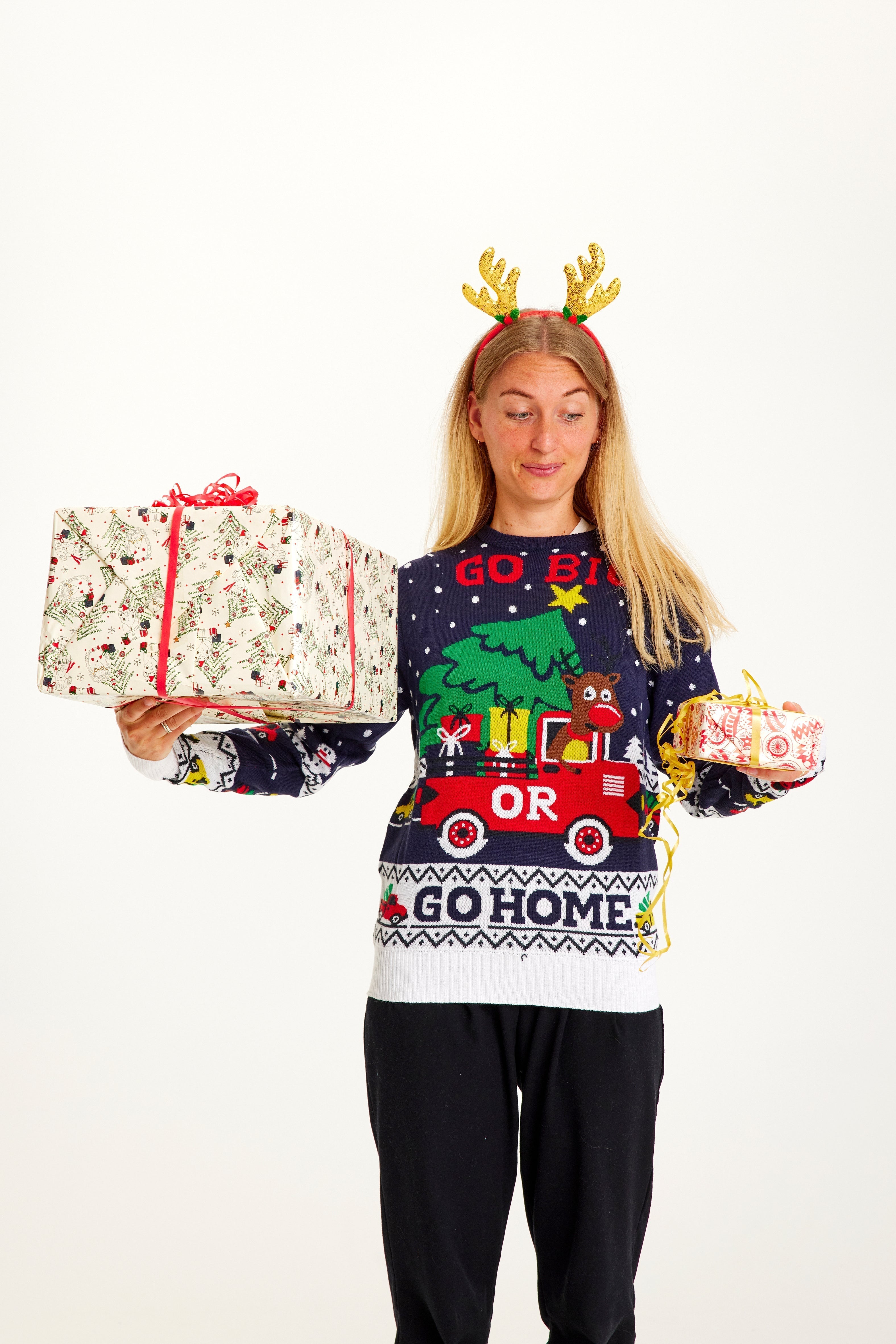 Une femme posant avec des cadeaux de Noël dans les mains et portant un pull de Noël avec la citation